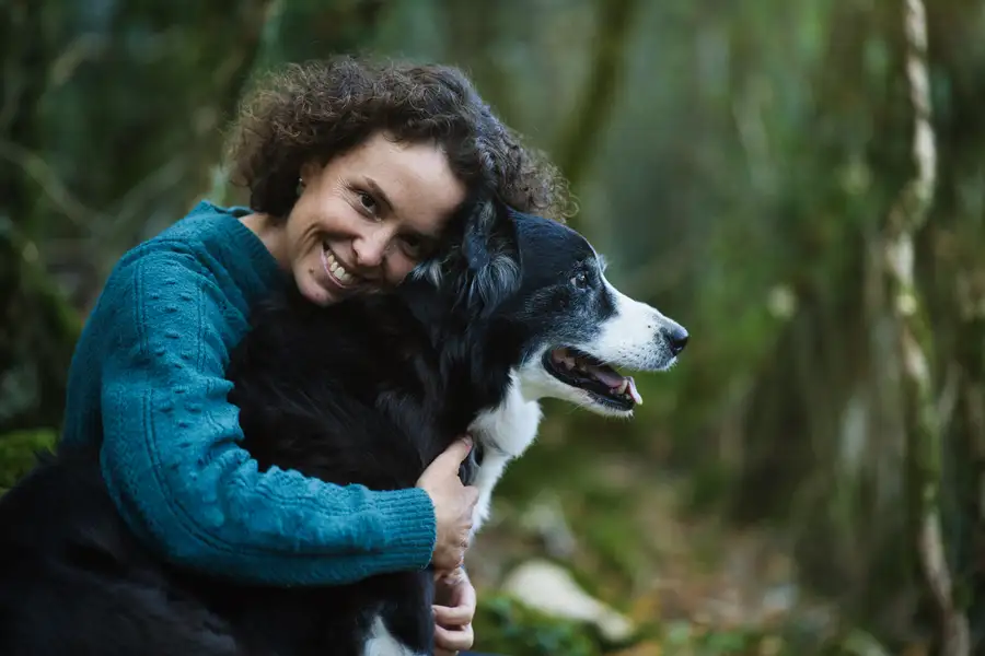 Une femme souriante serre contre elle son chien, un berger noir et blanc, en forêt